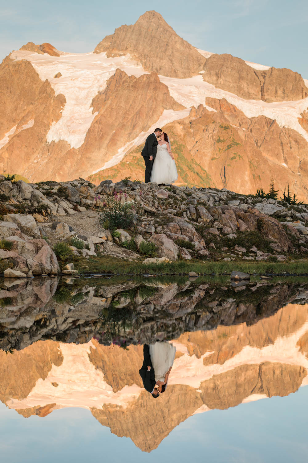 A couple in wedding attire hug with mountains and a lake