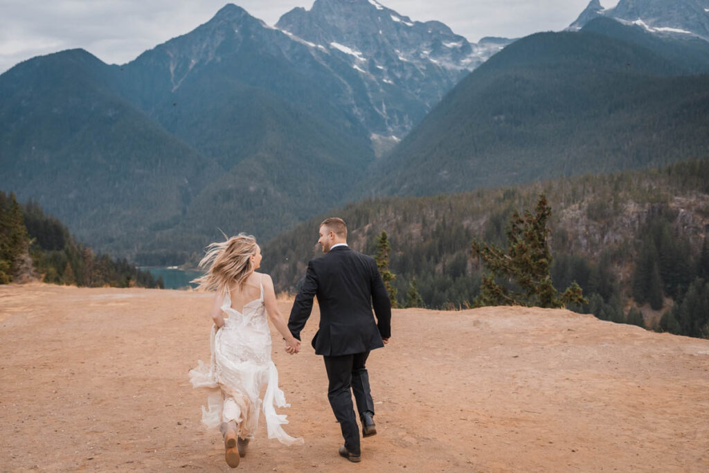 Two people run holding hands with mountains in the background