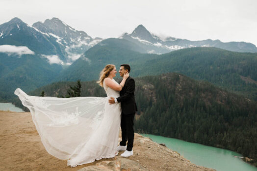 A couple in wedding attire hug with mountains and a lake in the background