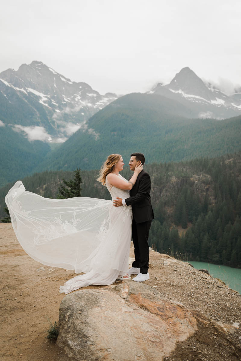 A couple in wedding attire hug with mountains and a lake in the background